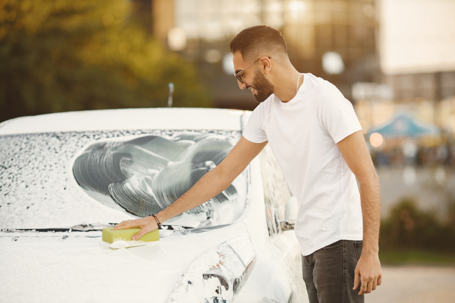 Young man in fashion clothes washing car at car wash station using washcloth. Man wearing white t-shirt and jeans. Indian man washing his white vehicle.